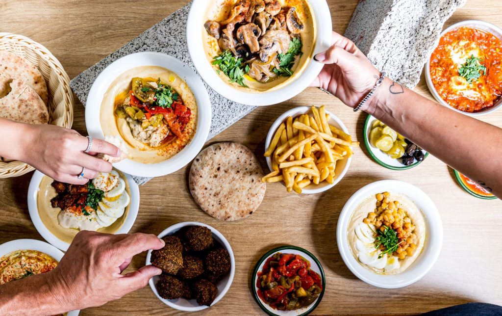 שולחן עם צלחות חומוס בתוספות שונות, קערת פלאפל, פיתות, צ'יפס וחמוצים, וידיים שמושיטות פיתה לחומוס